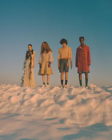 Four fashion models stand on a snowy hill under a clear sky. The models wear distinct outfits and pose for a fashion editorial.