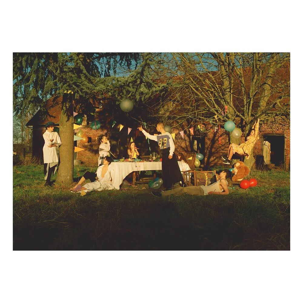 A group of seven young adults in an outdoor setting with a picnic table, balloons, and trees, set against a rustic building backdrop.