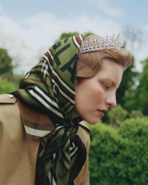 Blonde woman in trench coat and olive scarf wearing a tiara standing outside in natural light, fashion editorial style headshot.
