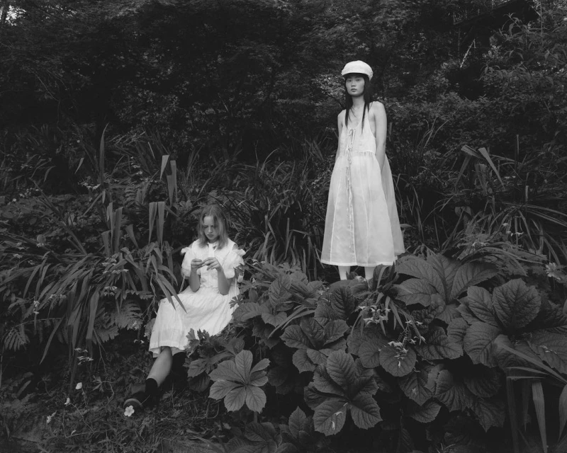 Monochrome image of two young women in white dresses and hats in a lush, overgrown garden setting with dense foliage and dark, moody lighting.