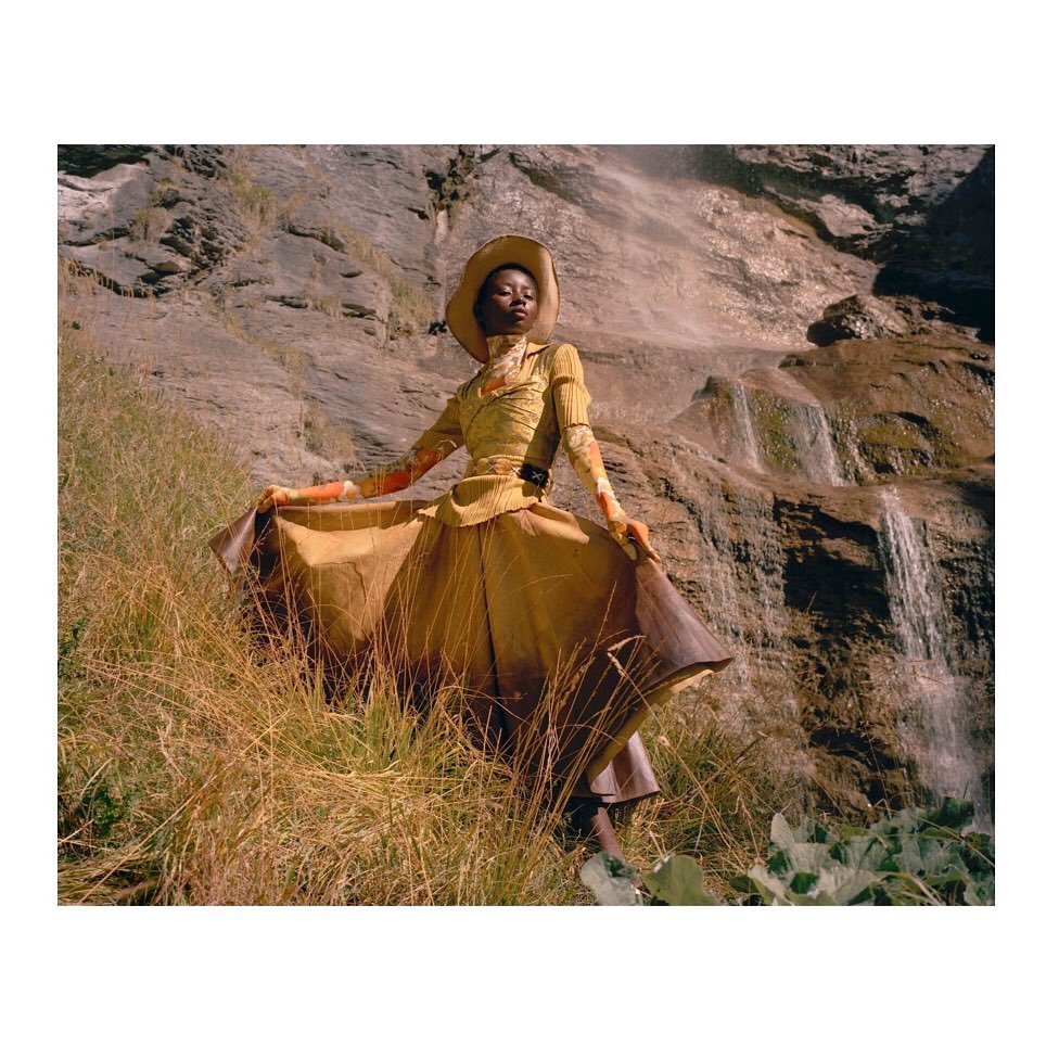 Young woman in yellow dress and hat poses in a grassy field with a rocky cliff and small waterfalls in the background, fashion shot.