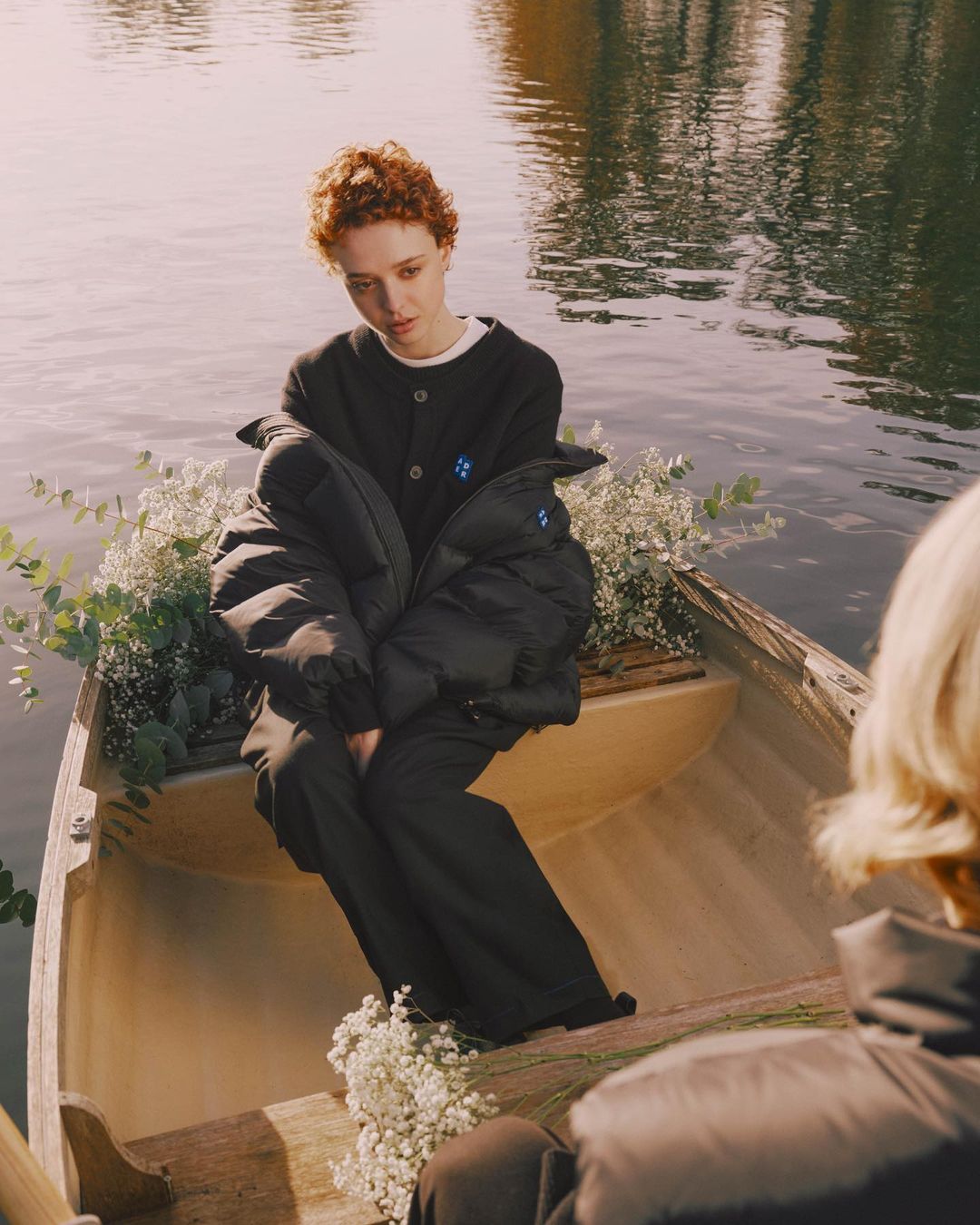 A woman in a rowboat surrounded by flowers, wearing a cardigan and jacket, floating on a lake in natural light, medium shot.