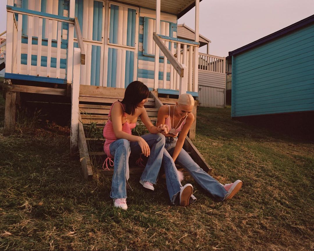 Two young women sit on steps of a blue-and-white beach hut, casually dressed in jeans and tops, enjoying a sunny, relaxed moment.
