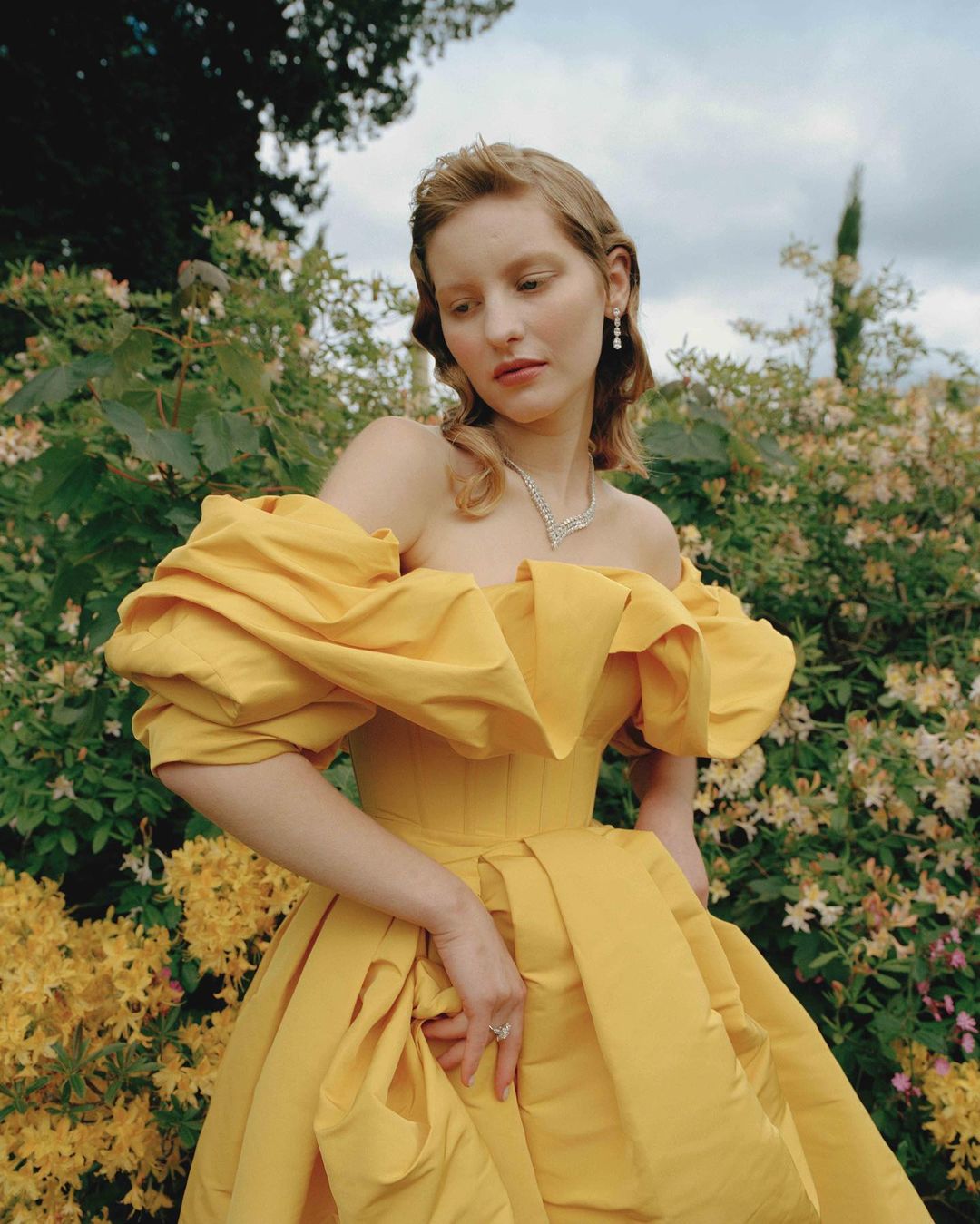 Woman in a yellow gown stands in a lush garden surrounded by green trees and yellow flowers in an eye-level outdoor shot.