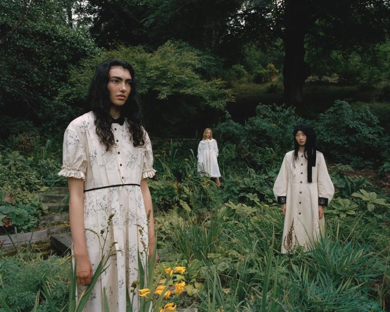 Three women stand in a verdant garden setting wearing light-colored dresses with a vintage, slightly gothic, fashion sensibility.