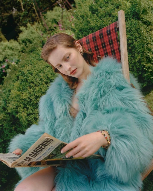 Woman in a turquoise fur coat relaxes outdoors, reading a gardening newspaper in a plaid deck chair, surrounded by lush green foliage.