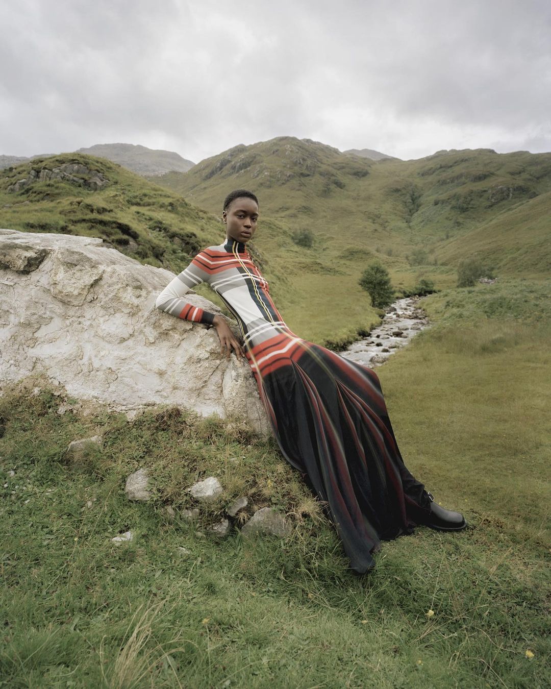 Woman posing in striped long-sleeve dress and maxi skirt leaning on rock in rural green hills with flowing creek under cloudy sky.