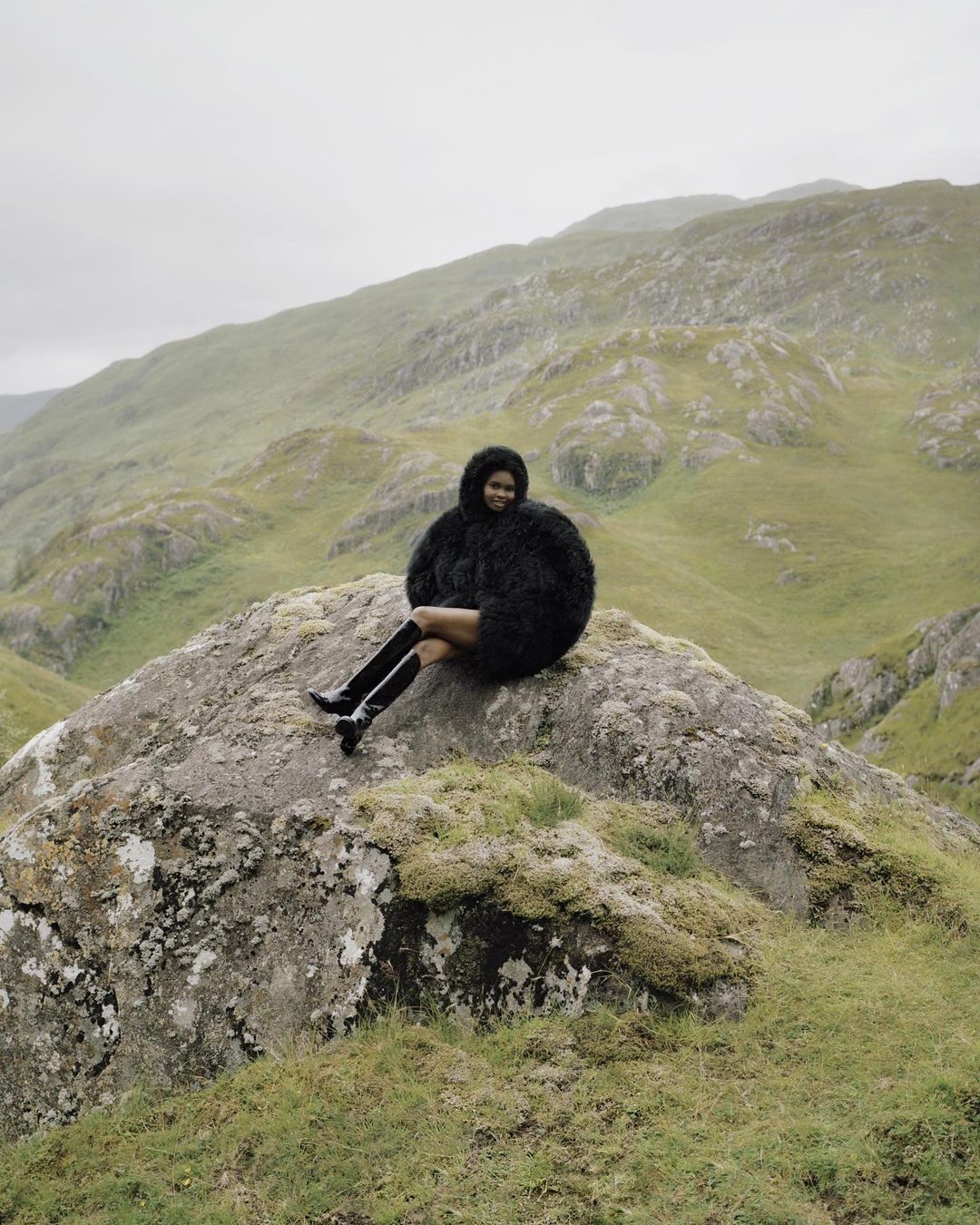 Woman in furry coat and boots sits on mossy rock in misty mountain landscape, gazing forward in an outdoor fashion setting.