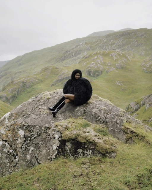 Woman in furry coat and boots sits on mossy rock in misty mountain landscape, gazing forward in an outdoor fashion setting.
