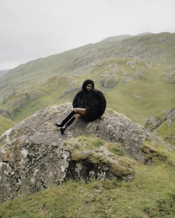 Woman in furry coat and boots sits on mossy rock in misty mountain landscape, gazing forward in an outdoor fashion setting.