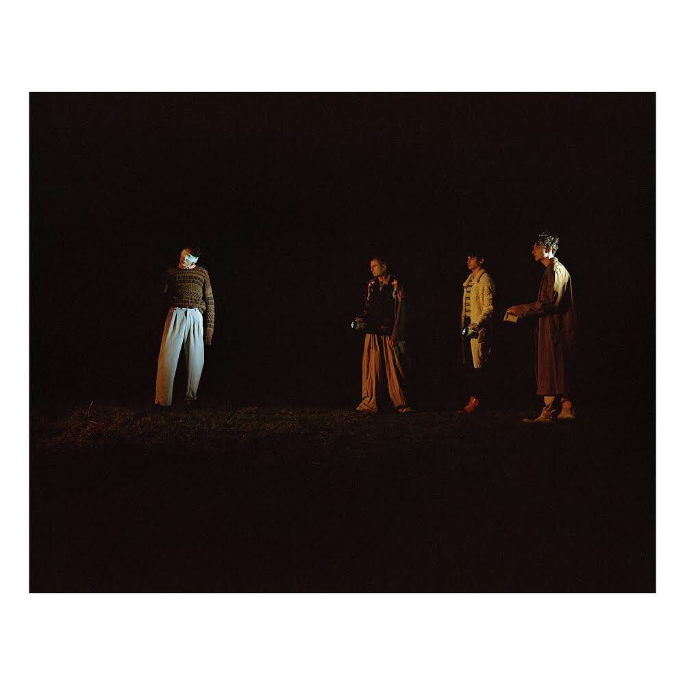 Four people stand in a dark outdoor space at night. One person faces away with white pants and a striped shirt.