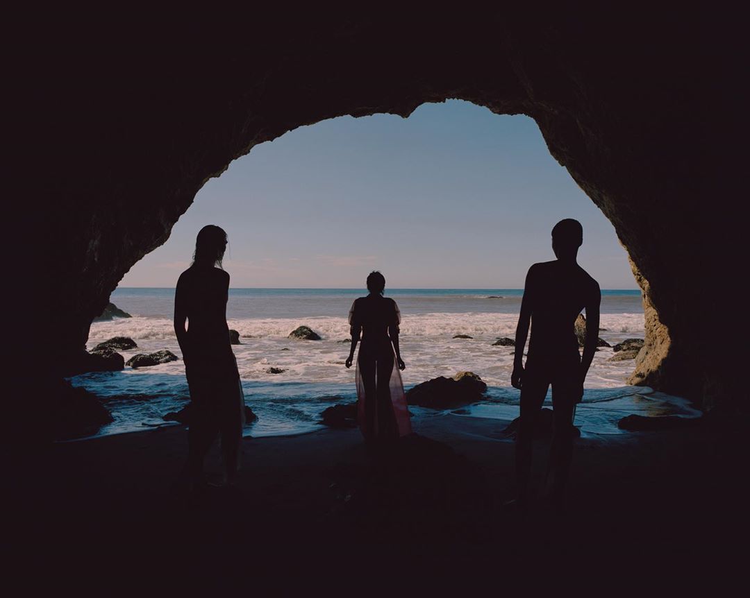 Three people silhouetted against ocean waves in a dark cave entrance, creating a striking contrast between light and shadow.