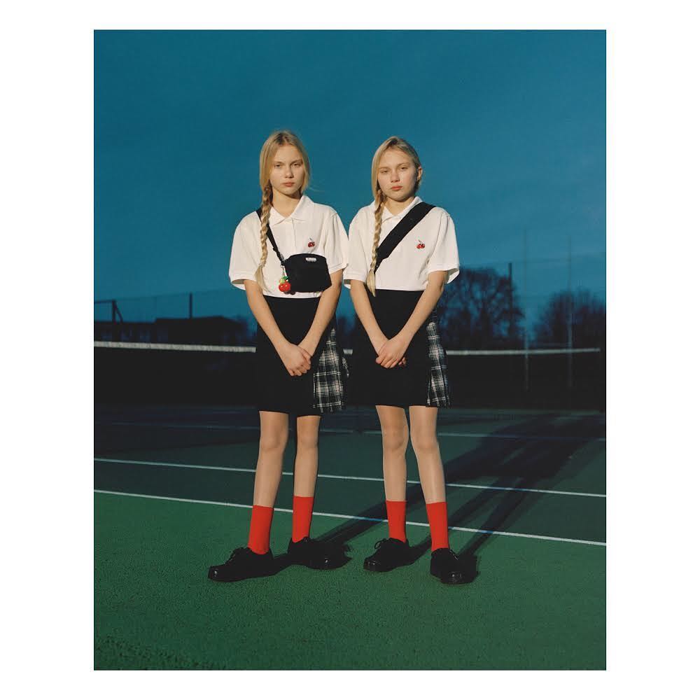 Two fair-skinned young women with braided hair wearing matching outfits, standing on a green tennis court against a blue sky background.