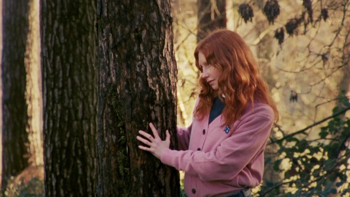A young woman with red hair wearing a pink cardigan touches a tree trunk in a forest setting, creating a peaceful, natural scene.