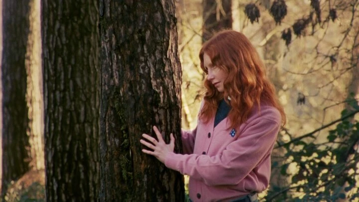 A young woman with red hair wearing a pink cardigan touches a tree trunk in a forest setting, creating a peaceful, natural scene.