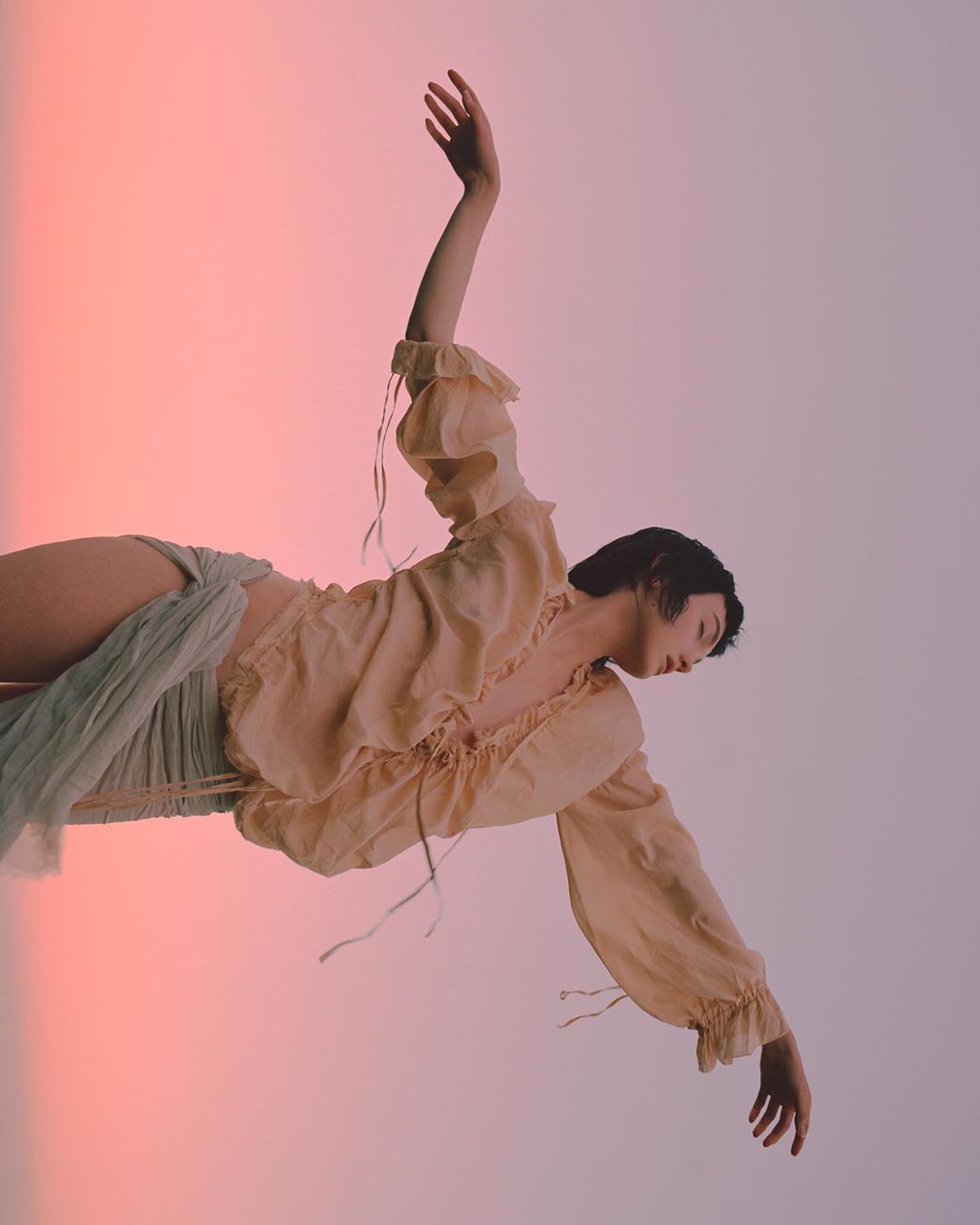 Female fashion model in a peach blouse and a white wrap posing in front of a gradient white and peach background in a studio shot.