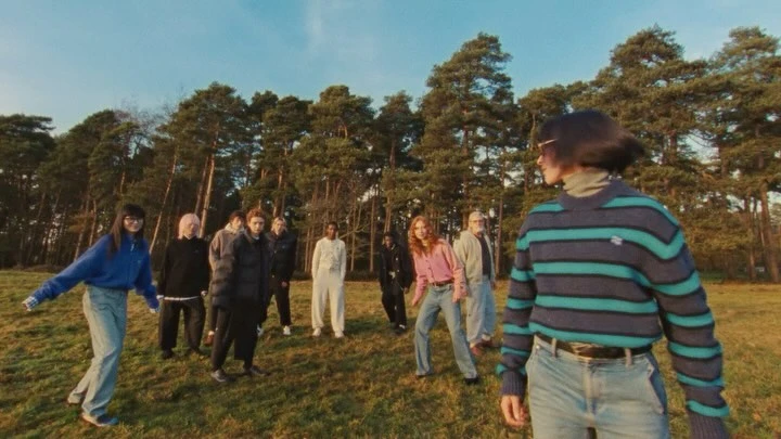 Group of nine people of mixed ethnicities standing on a grassy field with tall trees under a blue sky in daylight in a wide shot.