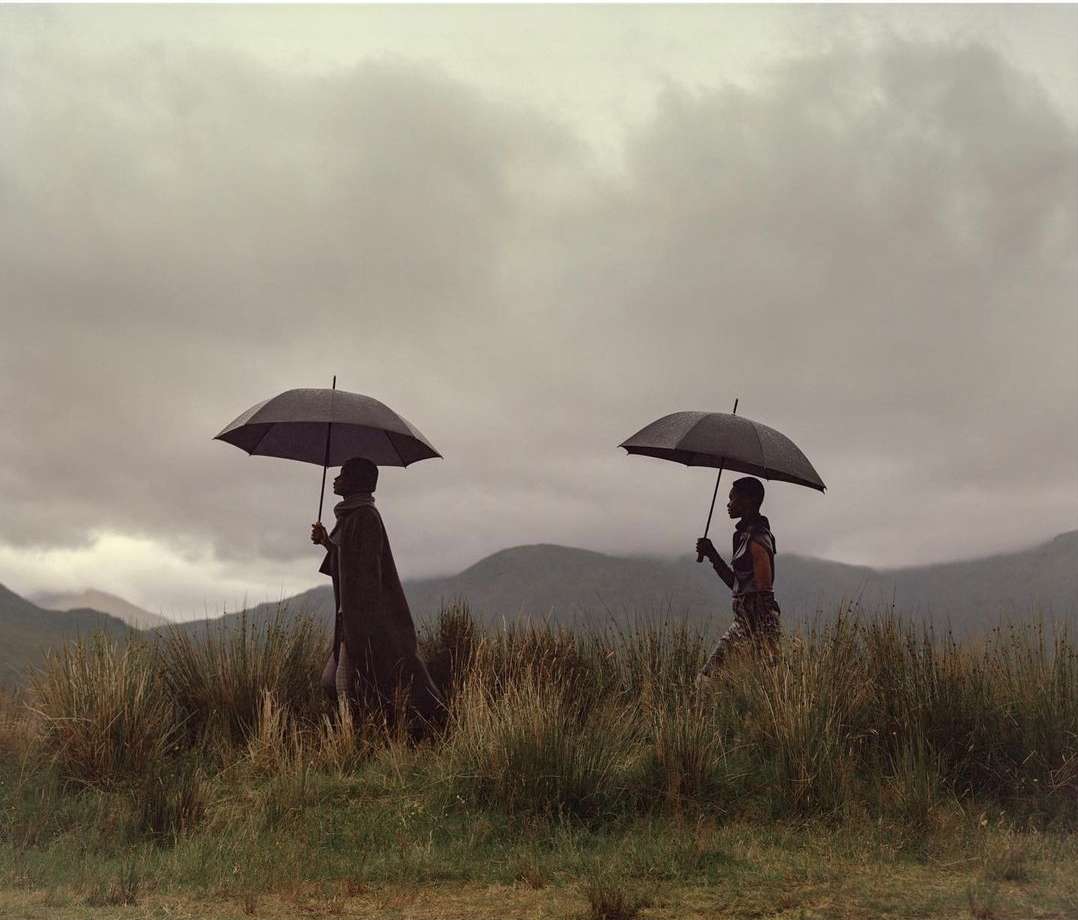 Two people in traditional clothing walk under umbrellas across a grassy field with mountains and cloudy sky in the background.