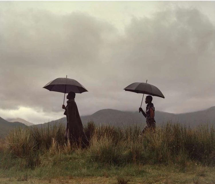 Two people in traditional clothing walk under umbrellas across a grassy field with mountains and cloudy sky in the background.