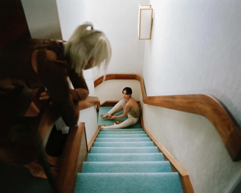 Two women pose on blue carpeted staircase. One woman sits and looks at camera, while second woman looks down from above.