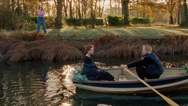 Two women are in a rowboat on a river with a third woman on the riverbank; trees and bushes line the background. 