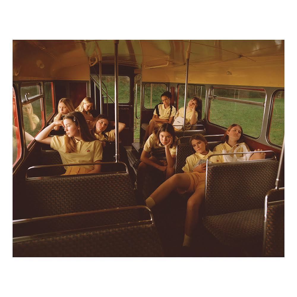 Group of pre-teen girls wearing yellow polo shirts inside a bus with brown interior and green scenery visible through the bus windows.