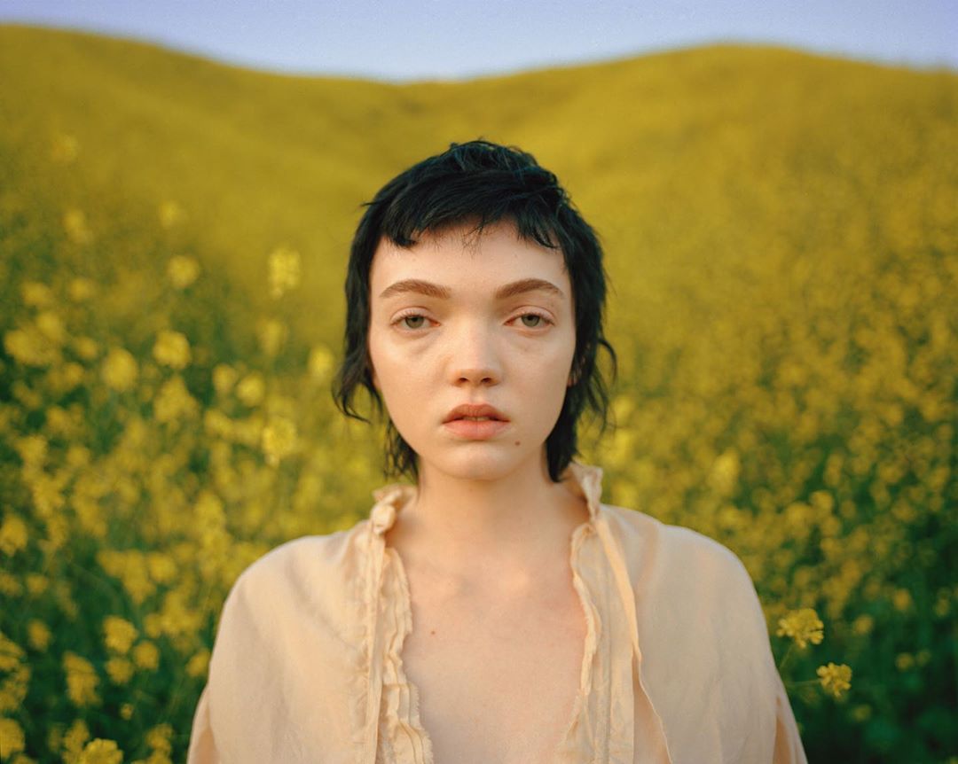 Close-up portrait of a young adult woman with short dark hair, gazing directly at the camera, set against a field of yellow wildflowers.