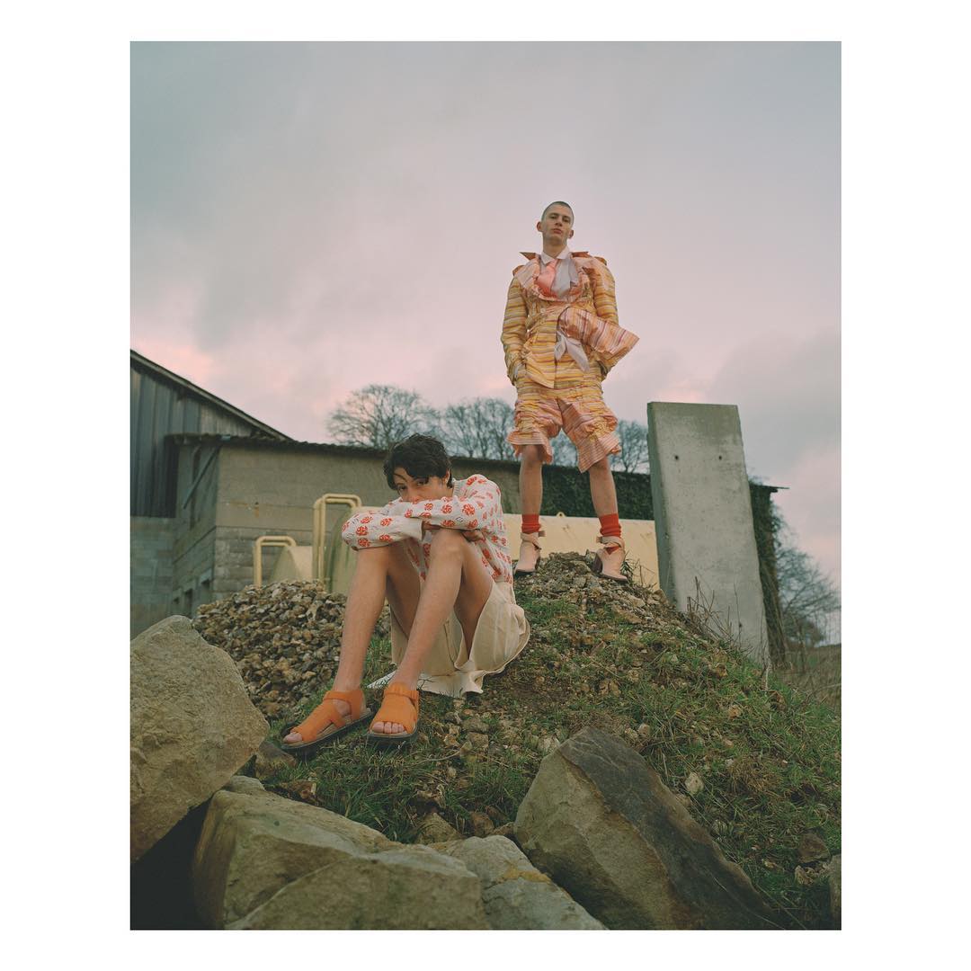 Two people are standing and sitting on a grassy hill in an outdoor setting, wearing uniquely designed fashion outfits on a cloudy day.