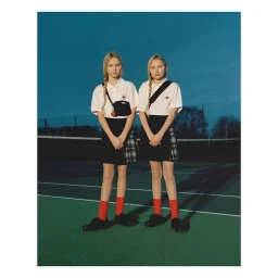 Two blonde girls in white polo shirts and dark skirts standing on a tennis court, photographed by James Perolls.