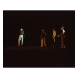 Four figures standing in a line outdoors at night, illuminated by direct light against a pitch-black background.