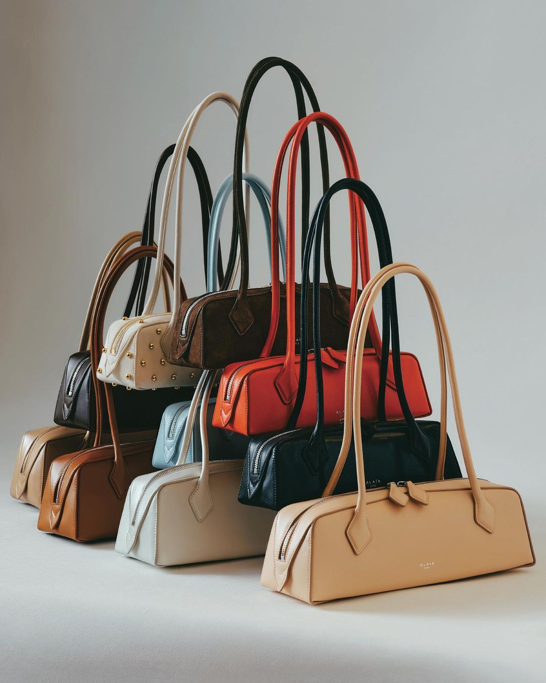 Collection of leather baguette bags in various colors, stacked against a plain background, showcasing fashion accessories.