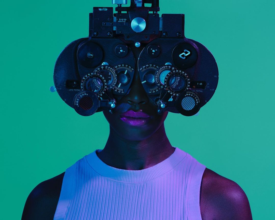 A person of color wearing an eye exam device against a green backdrop, bathed in teal and purple lighting for a modern, clinical feel.
