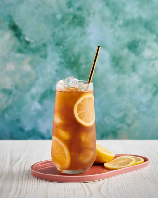 Iced tea in a glass with lemon slices and a gold straw on a pink tray against a teal background, evoking a refreshing beverage scene.