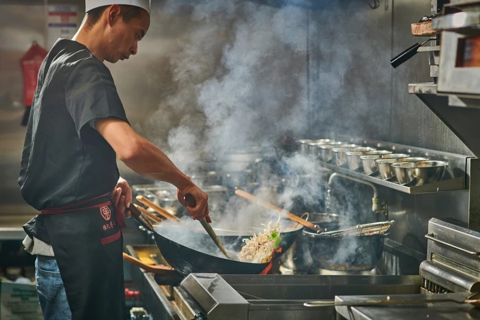 Professional chef stirring ingredients in a wok with high heat and smoke in a commercial kitchen setting, wearing a uniform.