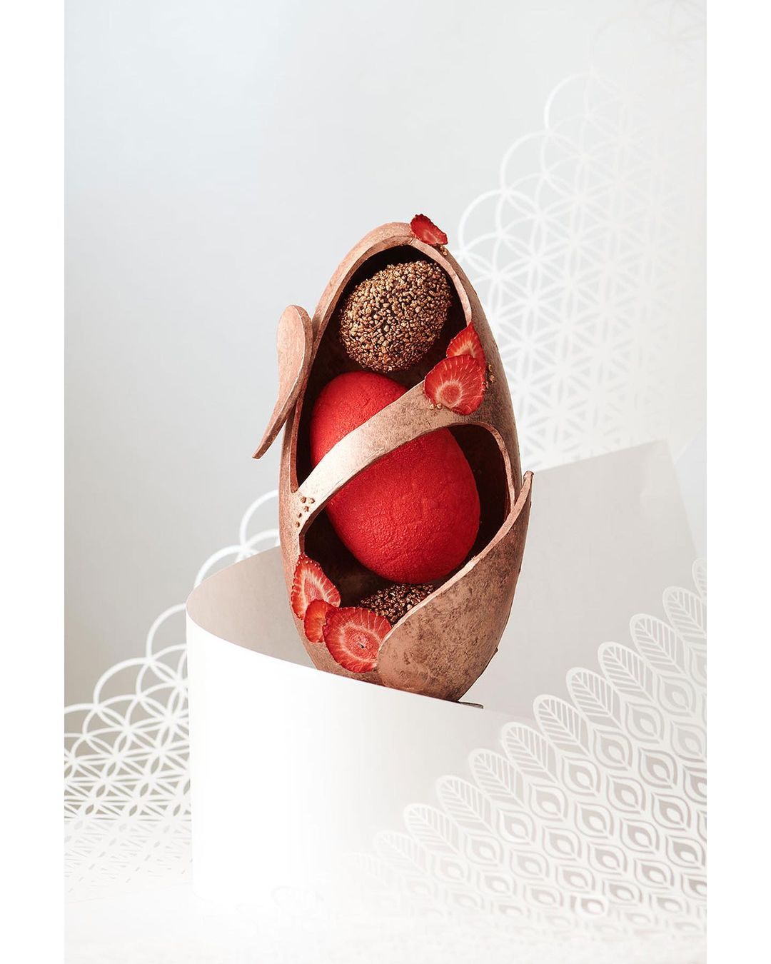 Sculpted chocolate Easter egg with red and brown spheres, garnished with fresh strawberries, on a white pedestal against a light background.
