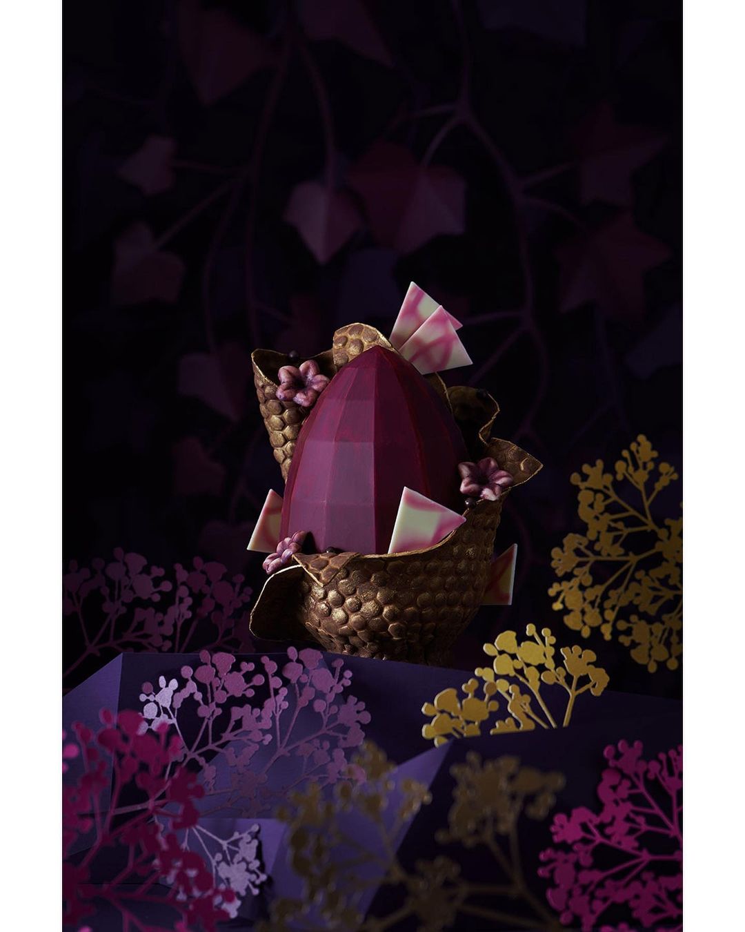 Geometric chocolate Easter egg sits inside a textured basket with floral accents against a dark, leafy backdrop. Festive dessert.