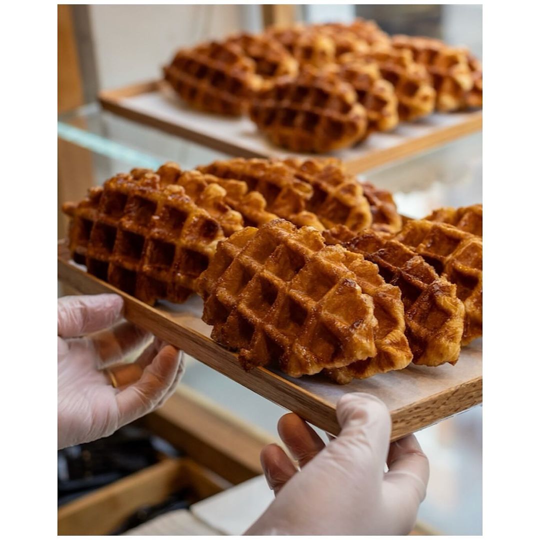 Close-up of golden-brown Liege waffles displayed on wooden trays, held by two hands wearing translucent gloves, food presentation.
