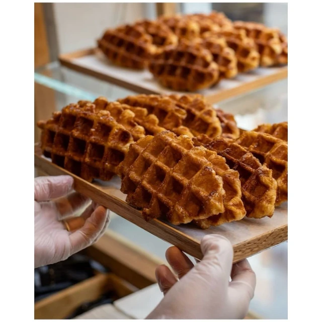 Close-up of golden-brown Liege waffles displayed on wooden trays, held by two hands wearing translucent gloves, food presentation.