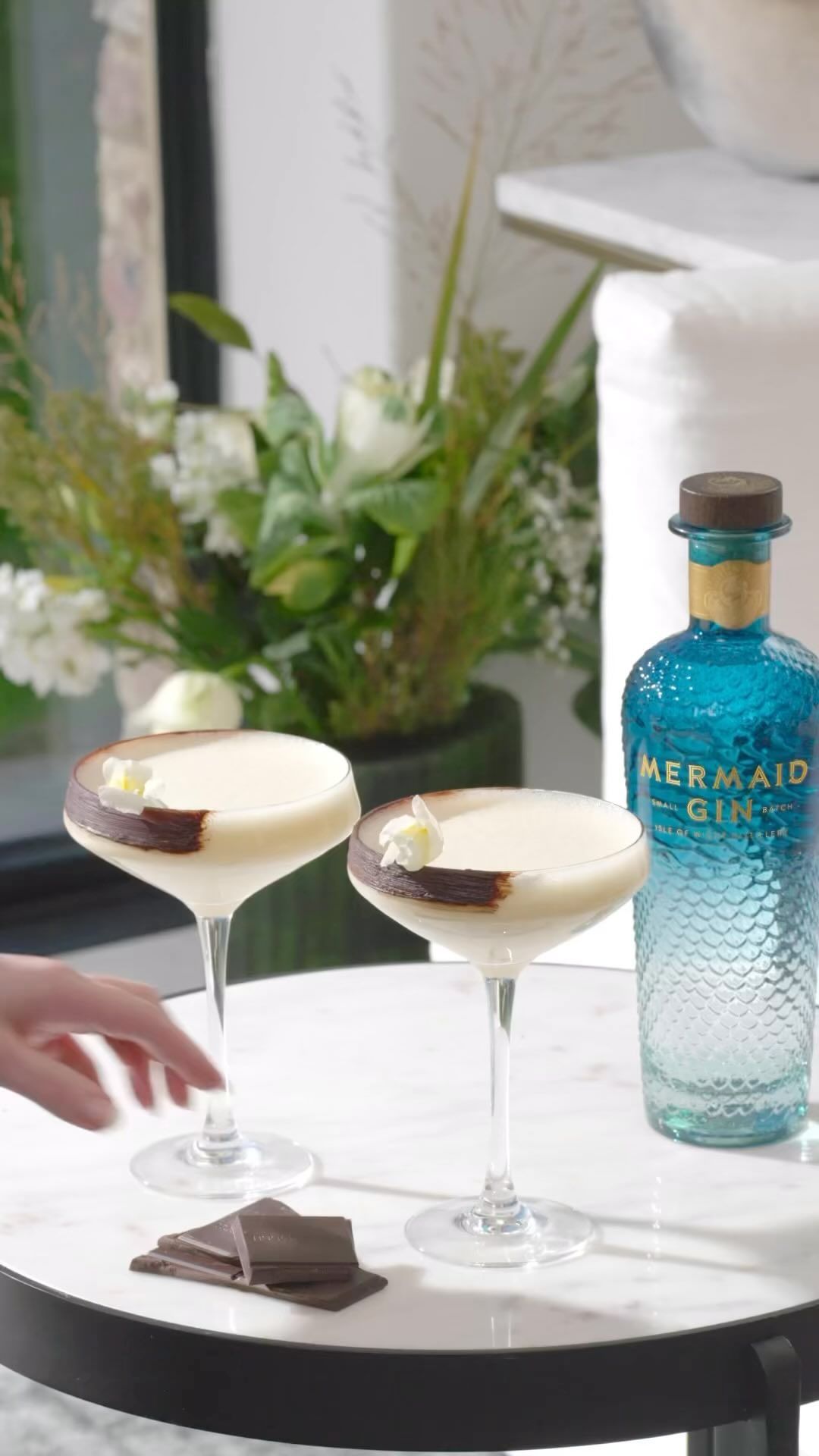 Two cocktails garnished with chocolate on a table with a bottle of gin and a floral arrangement in the background. Focus on drinks.