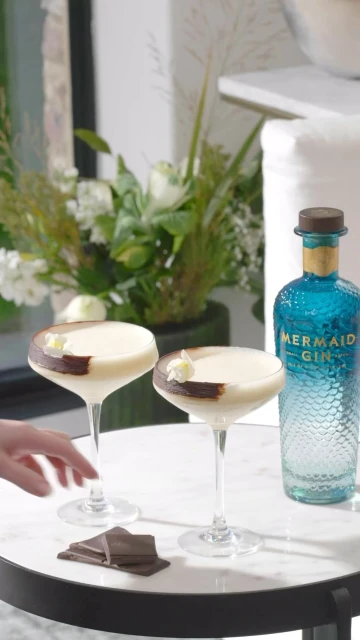 Two cocktails garnished with chocolate on a table with a bottle of gin and a floral arrangement in the background. Focus on drinks.