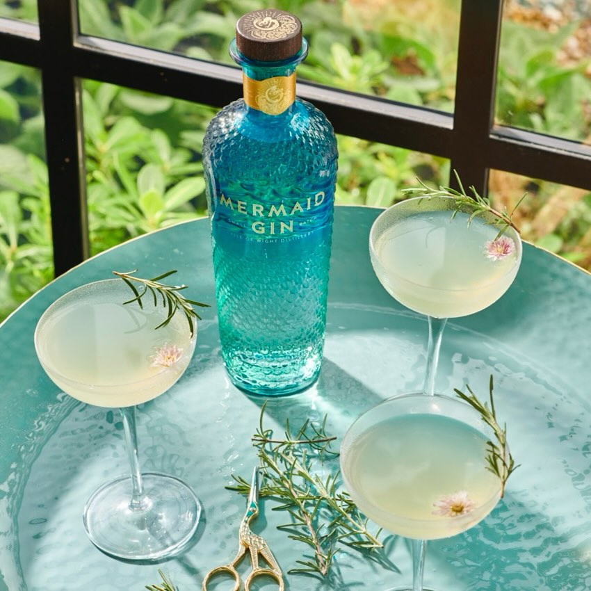 Mermaid Gin bottle displayed with stemmed cocktails and rosemary sprigs on a teal tray with a leafy green background.