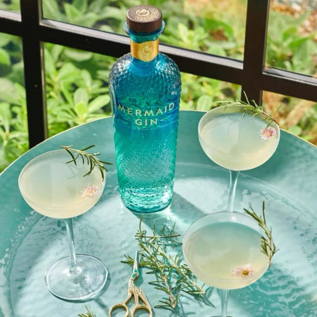 Mermaid Gin bottle displayed with stemmed cocktails and rosemary sprigs on a teal tray with a leafy green background.