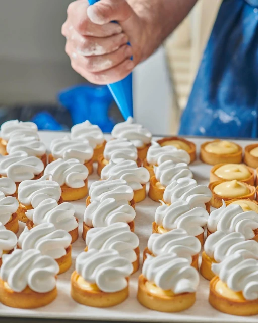 Close-up of a person decorating small tart pastries with white whipped cream using a piping bag, arranged in neat rows.