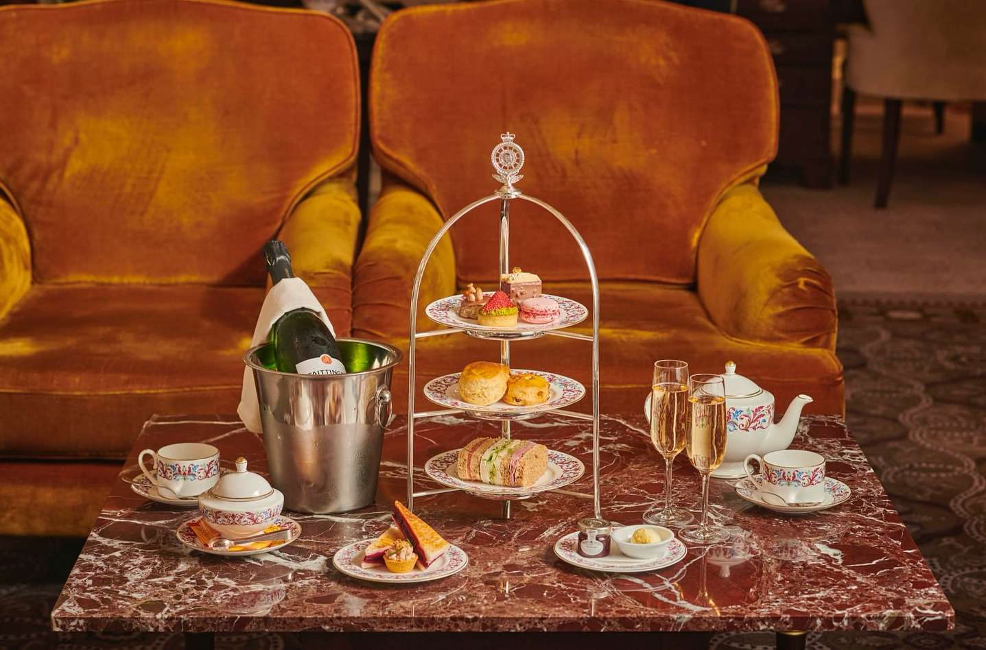 Champagne bottle in ice bucket with tiered dessert stand, teacups, and sandwiches on a marble table and two orange armchairs.