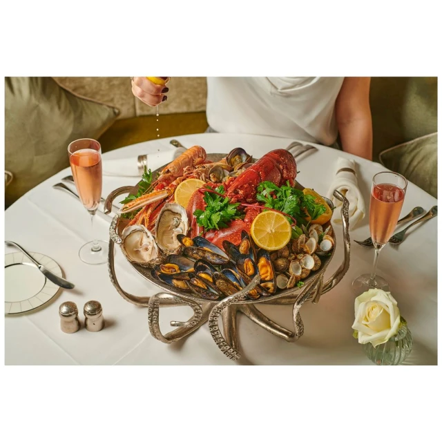 Seafood platter with lobster, oysters, mussels, clams on a silver octopus stand, sparkling wine, and a hand squeezing lemon juice.