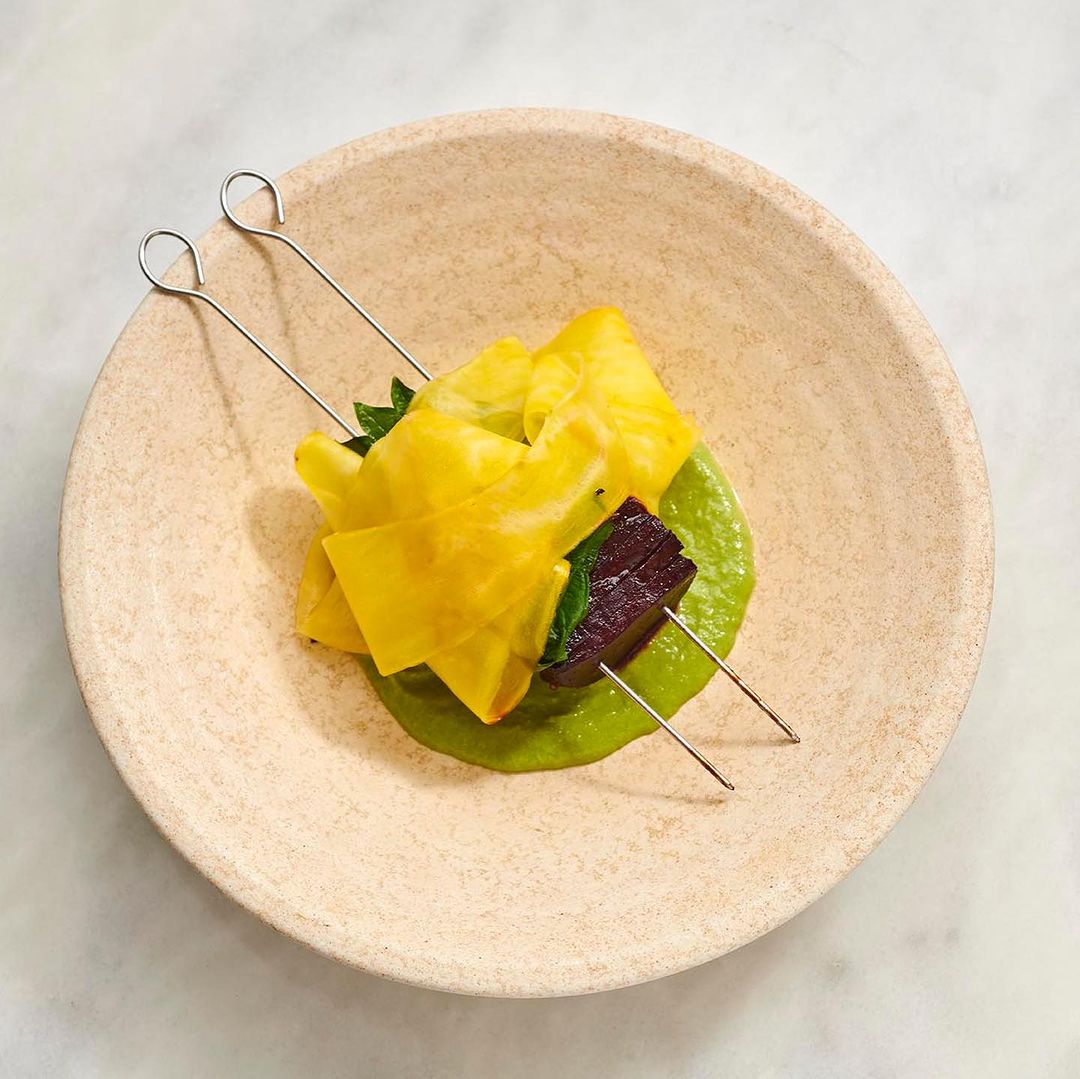 Gourmet food presentation of yellow fruit with purplish vegetable on a skewer over green sauce, on a tan plate, photographed overhead.
