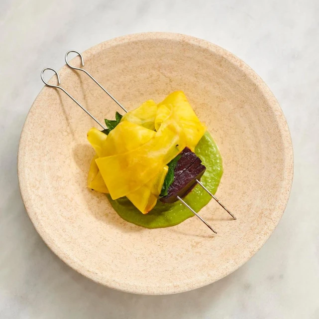 Gourmet food presentation of yellow fruit with purplish vegetable on a skewer over green sauce, on a tan plate, photographed overhead.