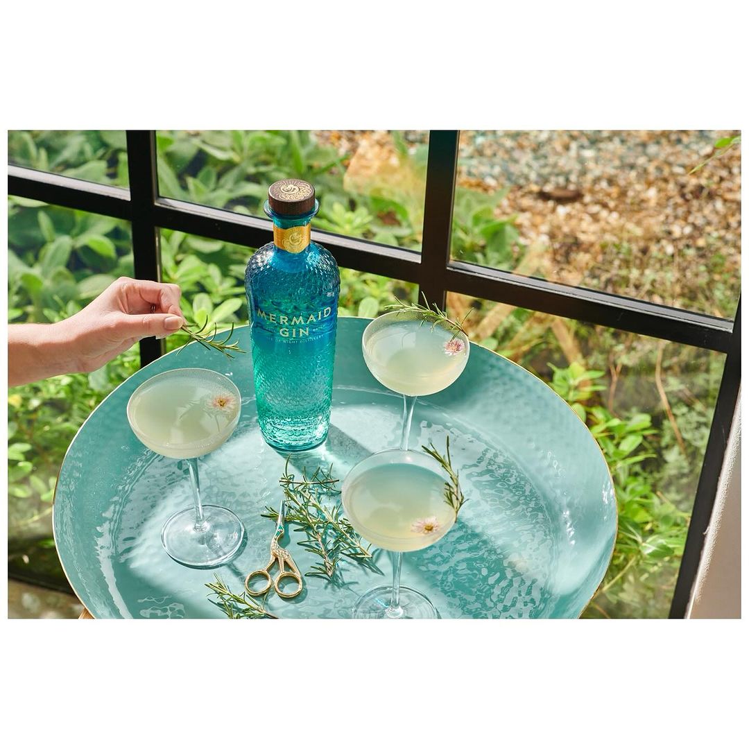 Mermaid Gin bottle and cocktail glasses filled with drinks and rosemary sprigs on a turquoise platter in front of a window.