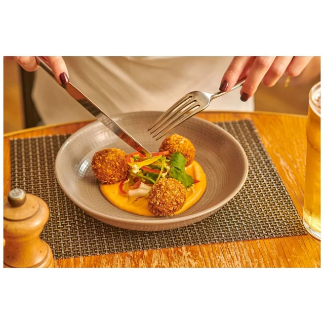 Overhead shot of an adult eating three breaded balls with a fork and knife on a grey plate at a wooden table in a restaurant.