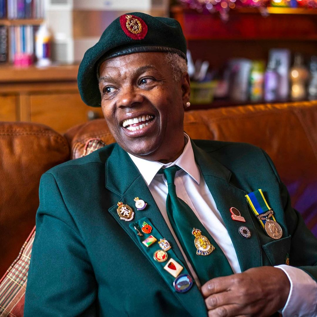 A smiling Black woman in a green military-style jacket and beret adorned with pins and medals indoors on a patterned sofa.